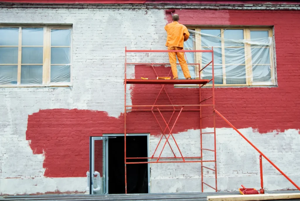 Man painting the outside of a commercial building red with a paint roller.