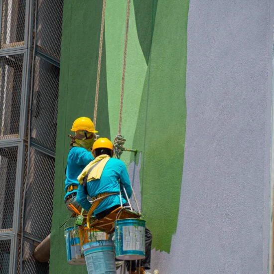 2 painters sitting on a suspended seat and painting the side of a building green.