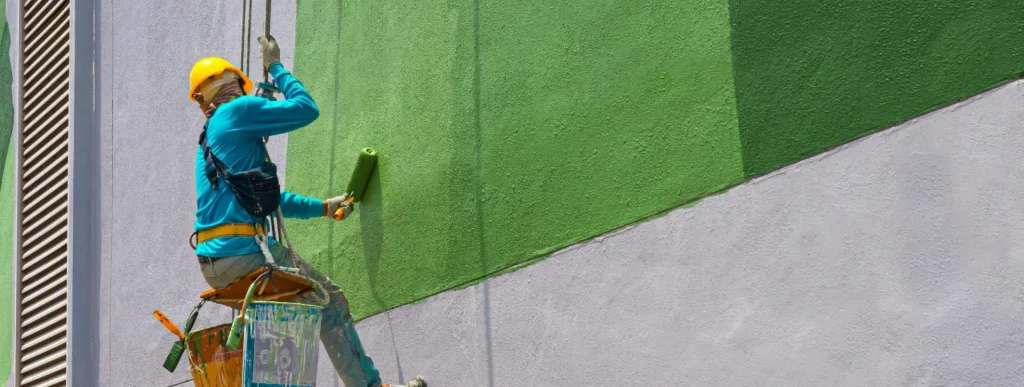 Man doing a painting service on the side of a building while sitting on a suspended seat.