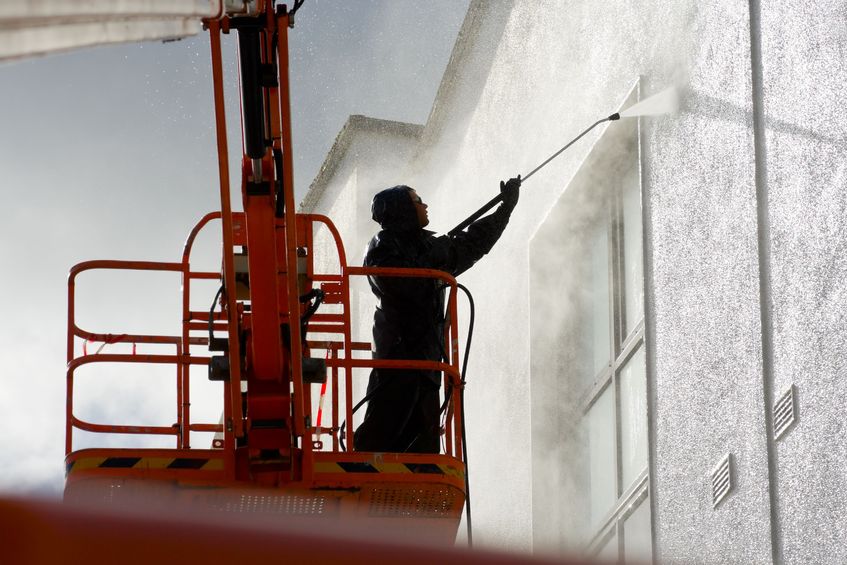 Worker using a pressure washer to clean the exterior wall of a tall building from a lift.