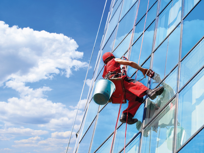 Professional window cleaner suspended by ropes, cleaning glass windows of a high-rise building under a clear blue sky.