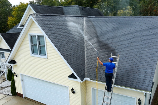 A professional on a ladder performing soft washing services on a house roof in Melbourne, ensuring safe and effective cleaning.