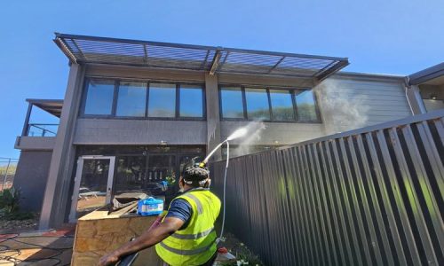 Worker performing building exterior cleaning with a pressure washer on a modern two-story building under a clear sky.